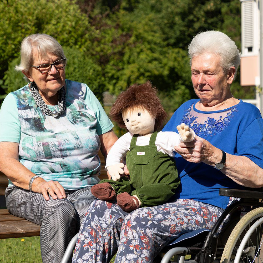 Zwei Seniorinnen beschäftigen sich mit der Joyk Empathiepuppe Marco im Garten
