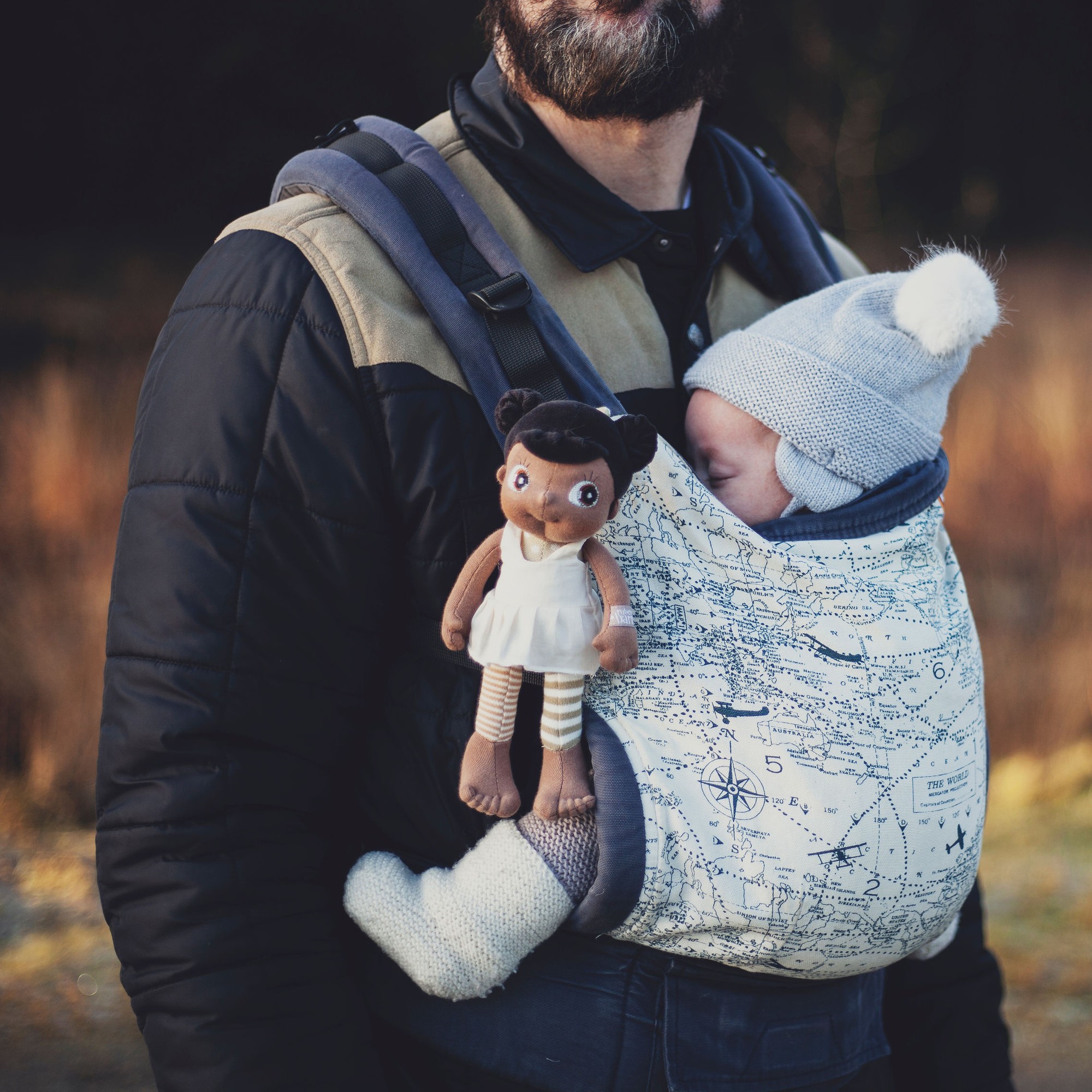 The Rubens EcoBuds mini doll Flora tucked into a baby carrier next to a baby
