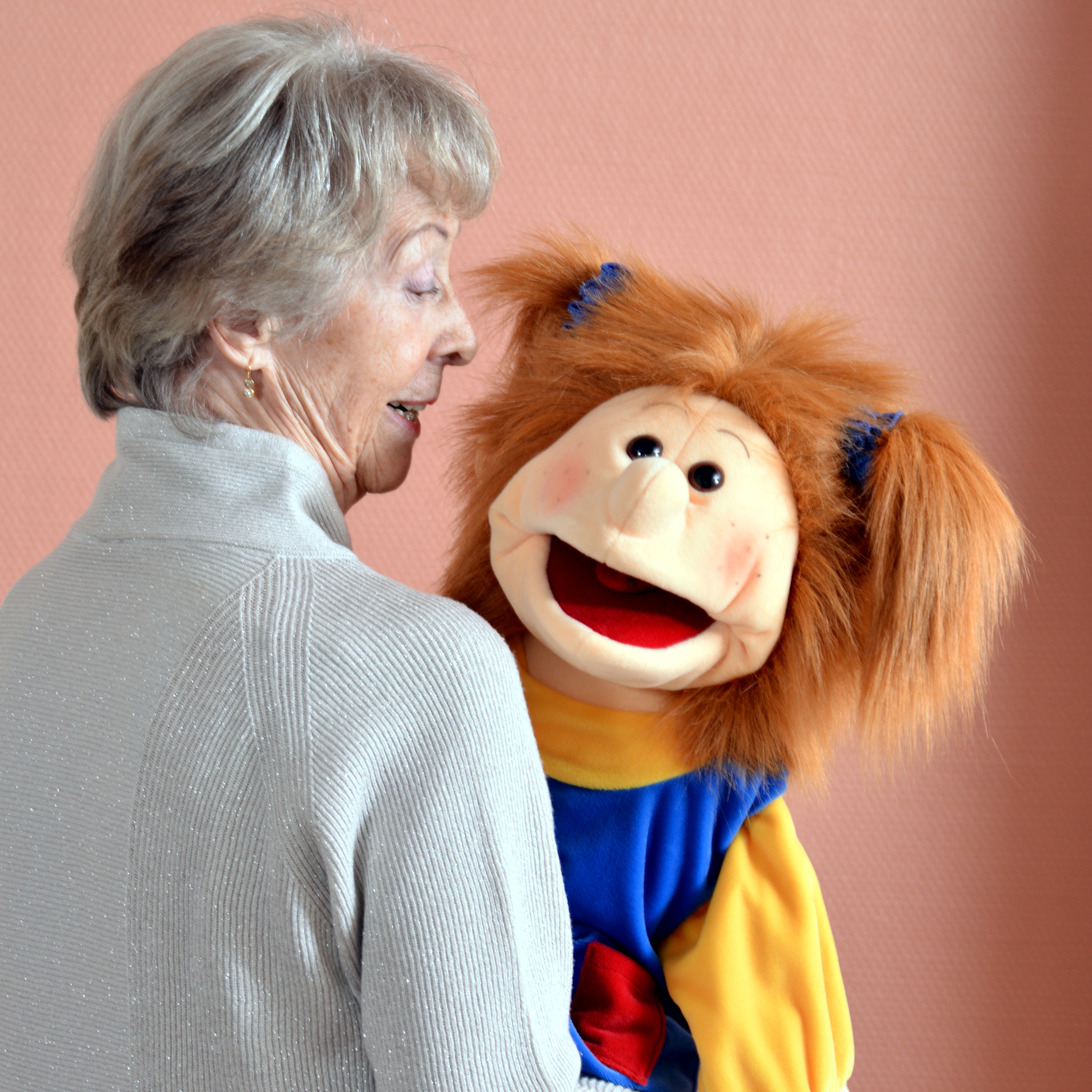Elderly woman holding the Living Puppets hand puppet Lotta with orange hair in her arms and looking at it, usage example