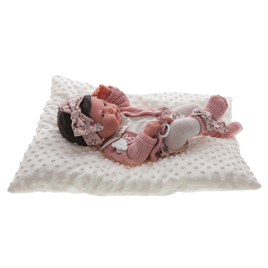 Antonio Juan baby doll Pipa, realistic doll with dark hair in a pink floral outfit with bow headband, lying on a white pillow