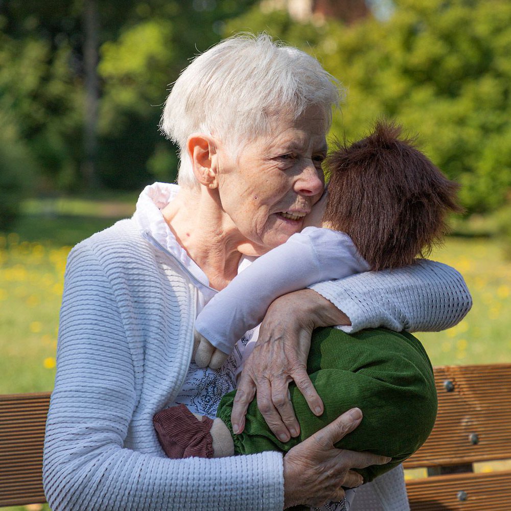 Seniorin umarmt die Joyk Empathiepuppe Marco auf einer Parkbank