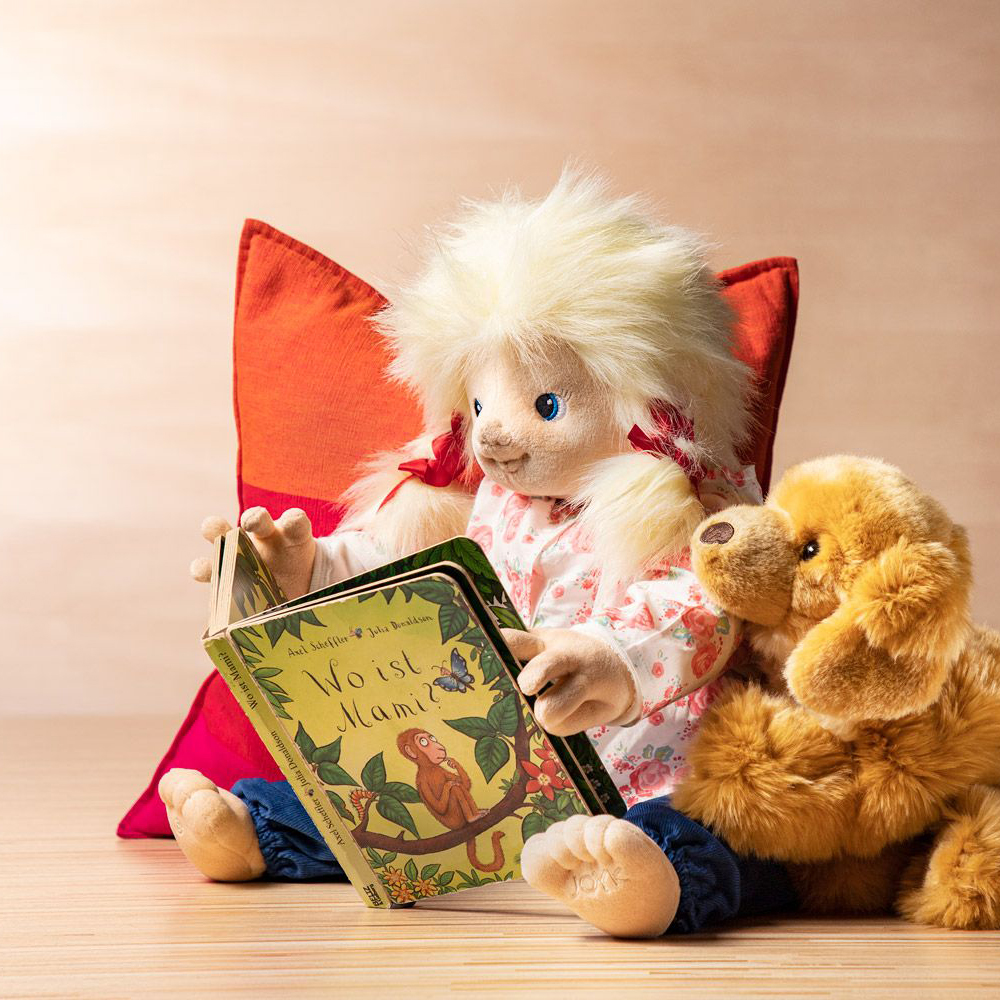 The Joyk empathy doll Nelly sitting with a picture book and plush dog against a cushion