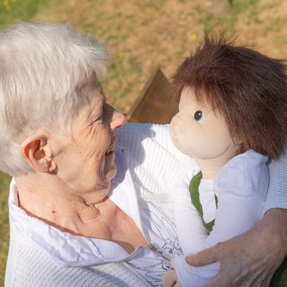 Seniorin im Blickkontakt mit der Joyk Empathiepuppe Marco im Garten