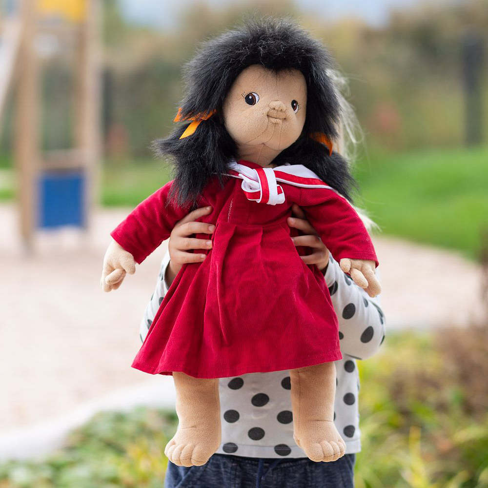 Kind trägt die Joyk Empathiepuppe Mia im roten Kleid auf dem Spielplatz