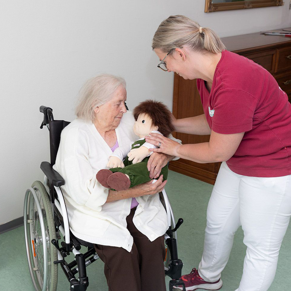 Caregiver handing the Joyk empathy doll Sam to a senior lady in a wheelchair