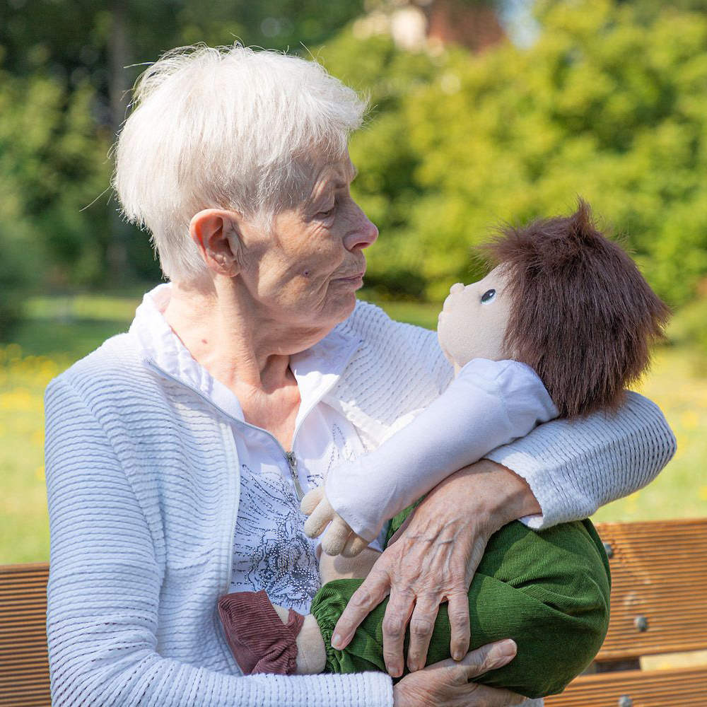 Seniorin hält die Joyk Empathiepuppe Marco im Arm auf einer Parkbank
